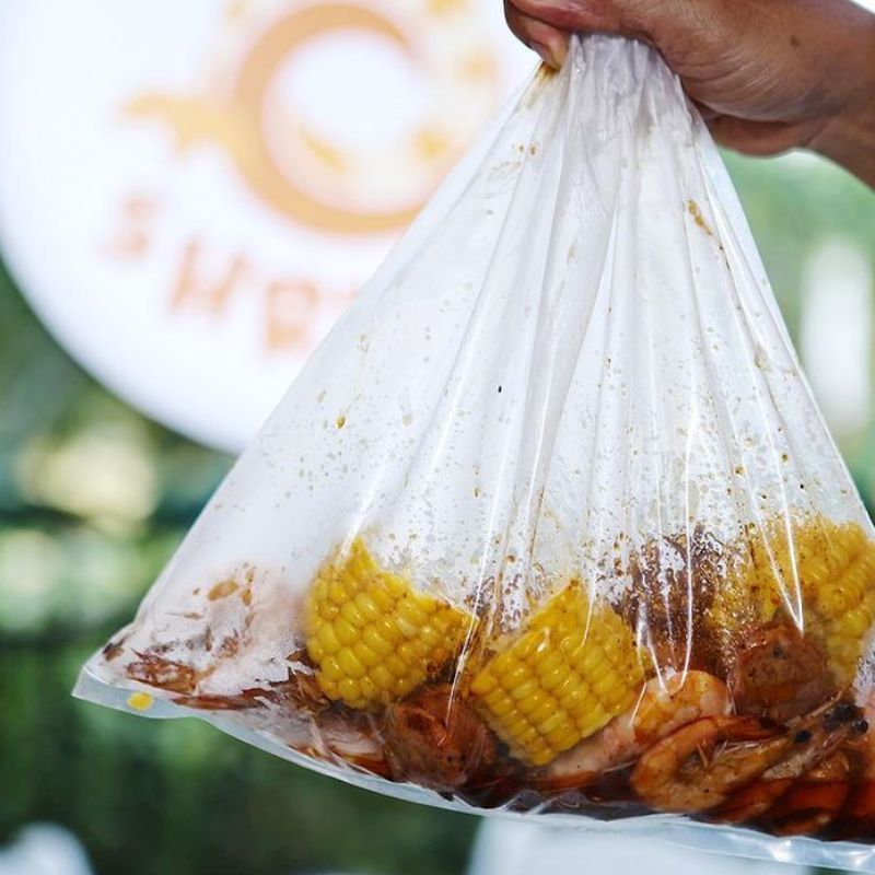 A hand holding up a sealed bag of shrimp and corn in front of the Bag O’ Shrimps sign