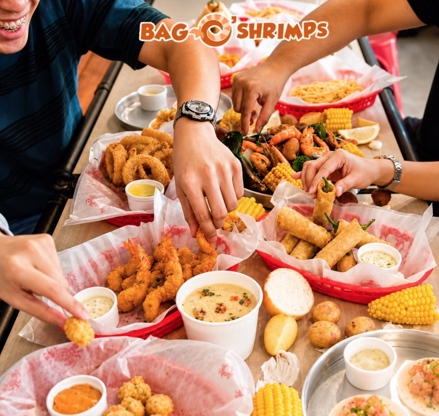 Friends reaching across a long table of fried shrimp, calamari, lumpia, chowder, corn and a seafood boil, with the Bag O’ Shrimps logo above